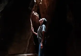 A girl standing in a cave looks up as light falls through a crack in the rock.