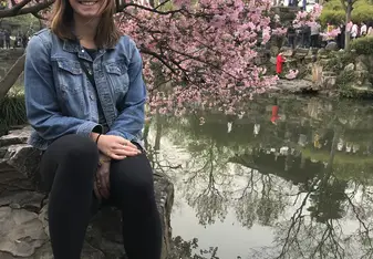 A young woman sitting on a rock with pink flowers behind her and a lake to the right