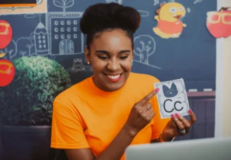 a woman in an orange shirt is smiling at a computer and pointing to a flashcard, which shows a picture of a cat and the letter C