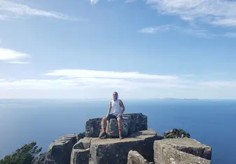 a young man in hiking clothes sits smiling on top of a rock overlooking the sea