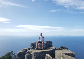 a young man in hiking clothes sits smiling on top of a rock overlooking the sea