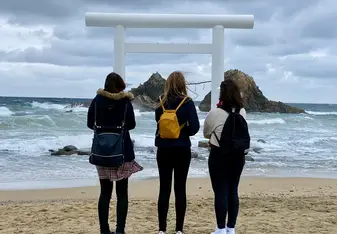 three girls standing on a beach with their backs to the camera, facing a large shrine in the ocean