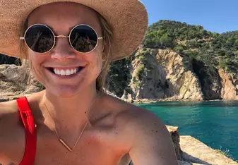 A smiling traveler wearing sunglasses and a sun hat takes a selfie in stunning Mallorca, Spain, with crystal-clear turquoise waters and rocky cliffs in the background. The perfect snapshot of teaching, exploring, and soaking up life with RVF International.
