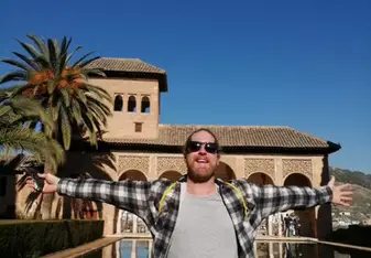Morgan stands with arms wide open, smiling in front of the stunning Alhambra in Granada, Spain. The clear blue sky and historic architecture reflect the joy and excitement of teaching and exploring Spain with RVF International.