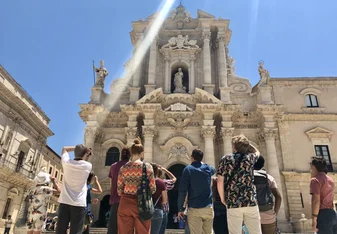 a group of students standing in front of Ortygia Cathedral, their backs are to the camera