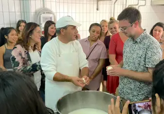 a group of students surrounding a man in all white demonstrating how to make cheese 