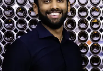 Vinay Vimalan wearing a navy button up shirt and a beard smiling