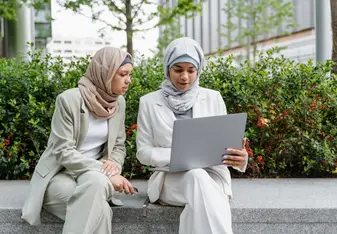 Two women with headscarves look at a computer screen outside.