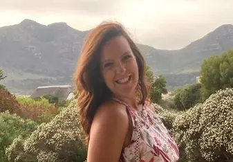 Tamsyn in a pink and red floral dress ,with red long hair smiling, and mountains in the background 