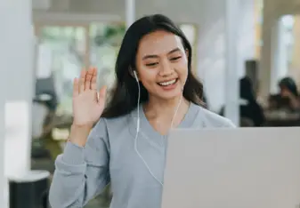 A person with brunette hair and headphones on waves at a computer camera
