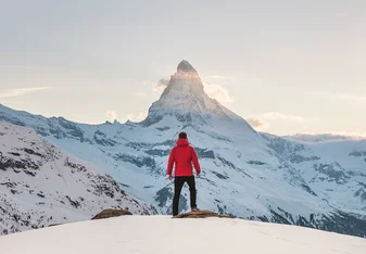A person with a red puffer jacket stands on a summit facing The Matterhorn mountain in the alps