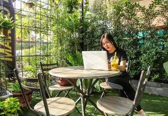 A woman looks at her laptop while sitting at a table surrounded by green plants.