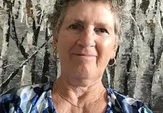 Jackie Bertrand Dix Jackson with short grey hair, earrings, and a blue geometric shirt smiling at the camera