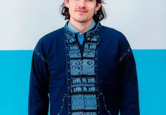William Davies wearing a blue traditional shirt and navy cardigan with long brown hair and blue eyes