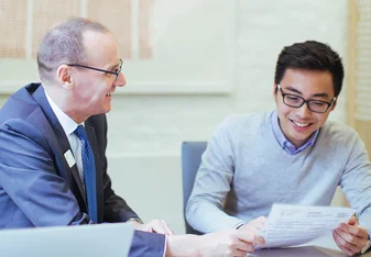 A male professor sits at a table next to a male student