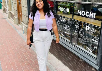 Kirsty Davids with white pants and a pink button up shirt and black hair smiling in front of a shop window