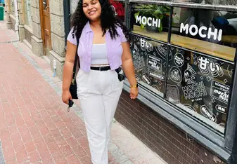 Kirsty Davids with white pants and a pink button up shirt and black hair smiling in front of a shop window