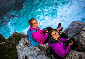 Two women sitting on a cliff by the ocean look up at and smile for the camera.