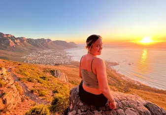 Amanda Drobek sitting at the top of a mountain looking over the ocean smiling with the sun setting