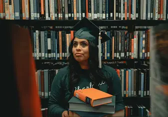 Student sitting next to bookshelf wearing graduation cap