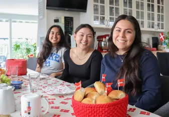 Three girls are sitting at a table and smiling. In front of them is a basket of baked good and Danish flags.