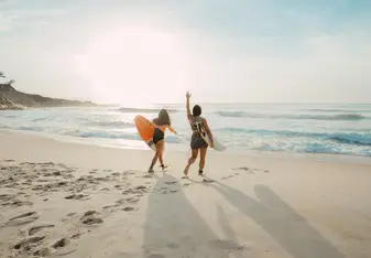 Two women holding surf boards walk on the beach.