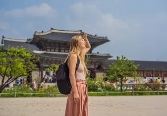 A woman wearing a backpack shields her eyes from the sun with a temple in the background.