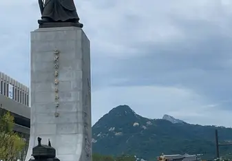 Statue of Admiral Yi Sun-sin in Gwanghwamun Square, Seoul, South Korea. The tall statue stands on a pedestal with Korean inscriptions, surrounded by a fountain and numerous visitors. The background features lush green mountains under a cloudy sky.
