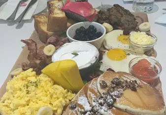 A large breakfast platter served on a wooden board, featuring scrambled eggs, pancakes with powdered sugar, sliced bananas, mango slices, bacon, sausage patties, dragon fruit, blueberries, French toast, and sunny-side-up eggs. Drinks including a mimosa and a glass of water with lemon are placed behind the platter, along with cutlery and condiments on the table.