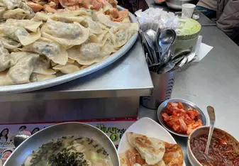 Street food stall serving a large tray of various dumplings with a bowl of noodle soup topped with seaweed, a plate of fried dumplings, and side dishes including kimchi and a spicy sauce. The stall is busy with customers, and the staff is actively preparing food in the background.