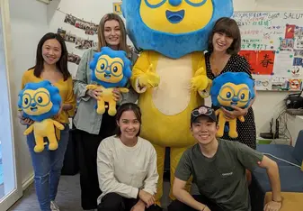 A group photo featuring five people and a large mascot in a room decorated with photos and posters. The mascot is a large, blue and yellow character with glasses and a cheerful expression. Four of the individuals are standing and holding smaller versions of the mascot, while two people are crouching in front. Everyone is smiling and appears to be happy. The room has white walls and a grey carpet, and the atmosphere seems friendly and joyful.