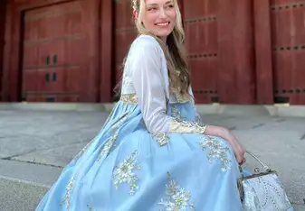 A woman dressed in a traditional light blue and white hanbok, adorned with intricate floral embroidery, sits gracefully on stone steps. She is smiling and looking off to the side, with a background of a traditional wooden building with red doors. The scene is peaceful and elegant, highlighting the beauty of the traditional attire and the historical setting.