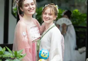 Two women in traditional attire, likely inspired by ancient East Asian fashion, are smiling and posing together in an outdoor setting with lush greenery. One is wearing a pink dress with elaborate hair accessories, and the other is in a white and green dress, holding a small blue and yellow plush toy. They stand close to a stone structure. The scene is vibrant and captures the beauty of cultural attire and natural surroundings.