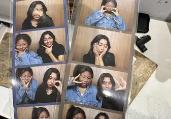 A close-up shot of two photo booth strips being held by someone with long, manicured nails. Each strip contains multiple small photos of two friends making various playful and expressive poses. The background of the photo booth appears to be wooden. One friend is wearing a denim jacket and glasses, while the other has long dark hair and is wearing a black top. The setting seems casual and fun, with the friends enjoying themselves in the photo booth. The strips are labeled "Mono mansion" at the bottom. In th