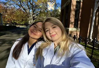  Two young women wearing white lab coats are standing outside on a sunny day, taking a selfie. They are smiling and look happy. Behind them, there is a large tree with bare branches and a building with brick walls. A decorative sign with a crest is also visible, indicating that they are likely on a university campus. The lab coats have a logo with the text "University of Otago" on them. The overall scene is bright and cheerful, capturing a moment of camaraderie and academic pride.