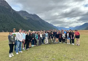 A large group of people posing for a photo in a picturesque outdoor setting. They are standing on a grassy field with majestic mountains and dense forests in the background under a cloudy sky. The group, consisting of men and women of various ages, appears to be enjoying an outdoor adventure or field trip. Everyone is dressed in casual, comfortable clothing suitable for hiking or exploring, and they are smiling and looking cheerful. The expansive natural landscape behind them adds to the sense of adventure 