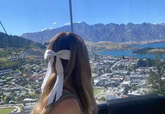A woman with long hair tied with a large white ribbon sits in a cable car, looking out at a stunning panoramic view. The scene includes a picturesque town, lush green hills, a sparkling lake, and rugged mountain peaks under a clear blue sky. The woman is wearing a light green sleeveless top and appears to be enjoying the beautiful scenery during her ride. The overall mood is serene and awe-inspiring, capturing the beauty of nature and the tranquility of the moment.