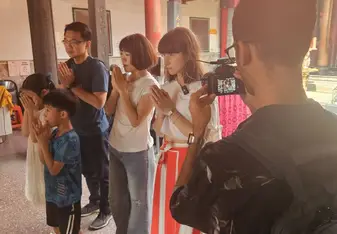  A group of people, including adults and children, are standing in a traditional temple with red pillars and ornate decorations. They are engaged in a prayer or meditation, with their hands clasped together in front of their chests. One person is recording the scene with a camera. The atmosphere is serene and respectful, reflecting the cultural and spiritual significance of the location.