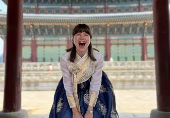 A woman dressed in a beautiful traditional hanbok, featuring a white and gold top with a dark blue skirt adorned with gold floral patterns. She is standing under an ornate, multi-colored roof structure and is laughing joyfully. She holds a small white and gold purse in front of her. The background shows the intricate architecture of a traditional building, enhancing the cultural richness of the scene. The woman's happy expression and the stunning surroundings create a lively and vibrant atmosphere.