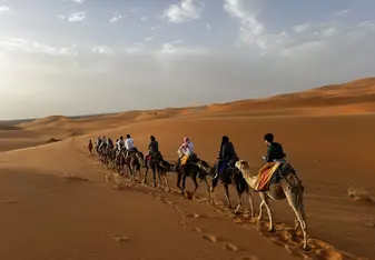 A group of people riding camels in a single file line across a vast desert landscape. The sand dunes stretch out into the distance under a partly cloudy sky, with the warm hues of the sand contrasting against the blue sky. The riders are dressed in a mix of traditional and modern attire, and they appear to be on a guided camel trek. The scene evokes a sense of adventure and exploration, capturing the beauty and tranquility of the desert environment.