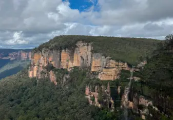 "A scenic view of a lush, green forested mountain range with sheer cliffs. The cliffs have a mix of greenery and exposed orange and brown rock faces. A waterfall cascades down the side of the cliff, blending into the dense forest below. The sky above is partly cloudy, with patches of blue sky peeking through."