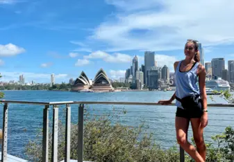 A woman stands on a walkway with a metal railing overlooking Sydney Harbour. She is wearing a light blue tank top, black shorts, and white sneakers. In the background, the iconic Sydney Opera House and the city skyline with its skyscrapers are visible across the water. The sky is clear and blue with a few scattered clouds