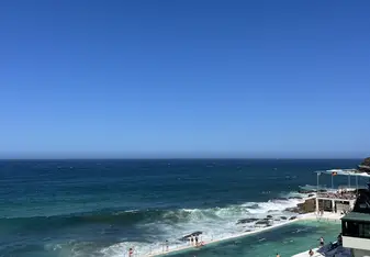 A coastal swimming pool filled with people enjoying the water on a sunny day. The pool is situated right next to the ocean, with waves crashing against the pool's outer walls. The ocean is a deep blue, and the sky above is clear and bright. The pool area is bustling with activity, with swimmers in the water and others lounging around the pool deck