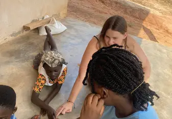 Four individuals are sitting on a concrete floor outside a building. Two local children and two volunteers, distinguishable by their attire and engagement, are interacting together. The children are wearing colorful clothing, while the volunteers, one in a light blue shirt and the other with light brown hair, are actively participating in a game or activity with the children, pointing their hands towards the center. 
