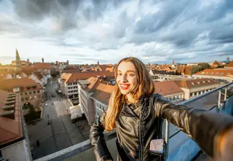 A woman smiles and takes a selfie on a rooftop with a German city in the background.
