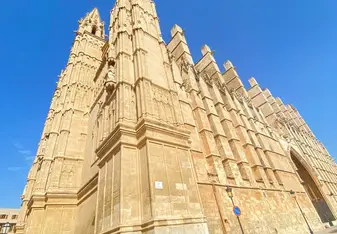A stunning view of a towering Gothic-style cathedral with intricate stone details set against a clear, vibrant blue sky. The grand structure's spires and architectural elements showcase its historic beauty in a sunlit plaza.