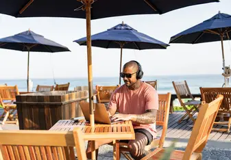 A man sitting outside under an umbrella works on a laptop.