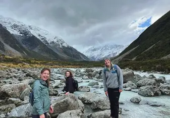 Three young women with backpacks stand on rocks and smile with mountains in the background.