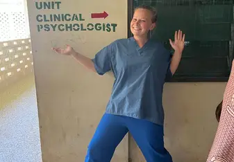 A volunteer in scrubs poses happily in front of a sign that reads 'Counselling Unit' and 'Clinical Psychologist'
