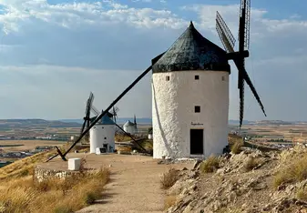 Traditional white windmills with black conical roofs sit atop a hill overlooking the plains of Castilla-La Mancha in Consuegra, Spain, under a blue sky with scattered clouds.